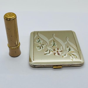 May include: Vintage gold-toned lipstick tube and a matching compact. The lipstick has a decorative gold band. The compact is square with a floral design in white, green, and pink. The compact has a gold clasp.