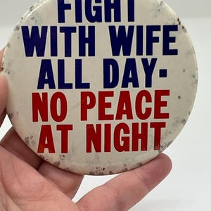May include: A circular, off-white badge with the words "FIGHT WITH WIFE ALL DAY - NO PEACE AT NIGHT" in blue and red. The badge has a slightly worn, vintage look.