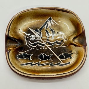 May include: Oval-shaped ceramic ashtray with a brown and tan glaze. The ashtray features a hand-drawn design of a boat with a mast, waves, and fish. The rim has two indentations for resting cigarettes.