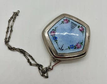 Op de afbeelding: Een zilverkleurige theedoos met een ketting. De doos heeft een vijfhoekige vorm met een lichtblauwe emaille oppervlak. Roze en blauwe bloemmotieven sieren het oppervlak. De ketting is aan de doos bevestigd.