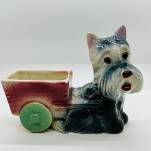 Può includere: Una fioriera in ceramica a forma di Scottish Terrier che tira un carro rosso. Il cane è principalmente grigio e bianco con occhi scuri e un naso nero. Il carro ha una ruota verde e un corpo rosso.