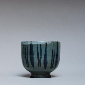 Puede incluir: Un cuenco de cerámica hecho a mano con forma redondeada y un esmalte moteado de color azul verdoso claro. Trazos verticales de pincel azul oscuro decoran el exterior. El cuenco tiene una base y un borde de color marrón oscuro y un interior azul más claro.