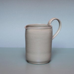 Puede incluir: Jarra de cerámica beige con asa curva. La jarra tiene un cuerpo cilíndrico y un pico para verter. El asa está unida al lateral. La jarra es de color neutro y tiene un acabado mate. Mide aproximadamente 15 cm de alto.