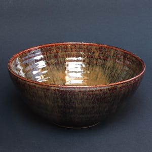 Puede incluir: Un cuenco de cerámica hecho a mano con exterior marrón oscuro y borde marrón rojizo. El interior tiene un esmalte brillante de color marrón dorado. El cuenco es redondo y tiene un diseño sencillo, adecuado para servir alimentos.
