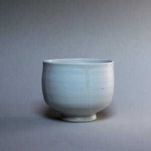 Puede incluir: Un cuenco de cerámica blanco roto hecho a mano, con un cuerpo redondeado y una pequeña base circular. El cuenco tiene un acabado liso y mate y una forma ligeramente irregular, lo que sugiere que está hecho a mano. El interior y el exterior son de un color gris azulado pálido.