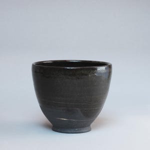 May include: A dark brown ceramic bowl with a glossy finish. The bowl has a rounded shape with a slightly flared rim and a small, circular base. The bowl is approximately 4 inches tall and 6 inches in diameter.