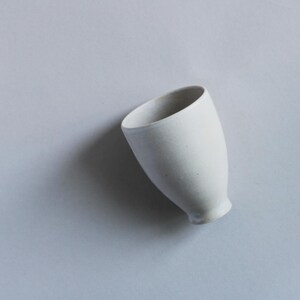 Puede incluir: Una pequeña taza de cerámica blanquecina con un borde ligeramente inclinado. La taza tiene un cuerpo redondeado y una base pequeña y acampanada. El interior es blanco y el exterior tiene un acabado mate. La taza está sobre un fondo gris claro.