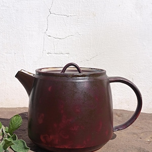 Puede incluir: Una tetera de cerámica hecha a mano con un esmalte burdeos oscuro. La tetera tiene un cuerpo redondeado, un asa curva y un pico. La tapa tiene un pequeño asa redondeada. La tetera está sobre una superficie de piedra.