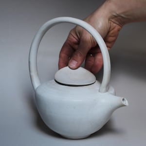 Puede incluir: Una tetera de cerámica blanca con un asa grande y arqueada y una tapa a juego. La tetera tiene un cuerpo redondeado y un pico. La imagen muestra la tetera sostenida por una mano.
