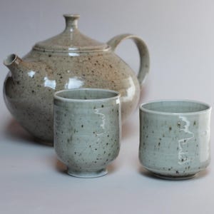 Può includere: Una teiera in ceramica con coperchio e manico, accompagnata da due tazze cilindriche abbinate. Il set presenta uno smalto color beige chiaro e grigio con una consistenza maculata su tutta la superficie della teiera e dei due piccoli contenitori.