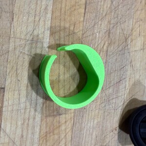 Puede incluir: Objeto circular verde brillante con una abertura curva. El objeto parece estar hecho de un material blando. Una parte de un objeto circular negro es visible en la esquina inferior derecha. El fondo es una superficie de madera marrón claro.
