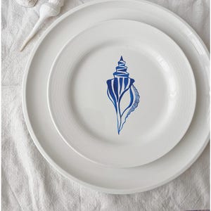 Pode incluir: Um prato de cerâmica branco com um desenho de concha azul. O prato está aninhado dentro de um prato branco maior.