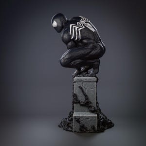 May include: A black and white statue of a crouching superhero, with a white spider emblem on the back. The figure is on a gray, textured pedestal with dark, organic details. The statue has a detailed, muscular physique.