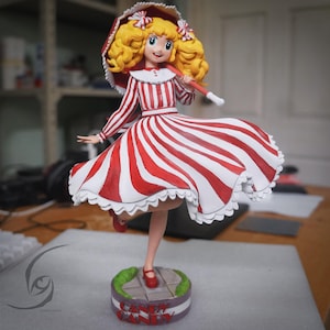 以下が含まれることがあります： キャンディキャンディのフィギュア。金髪の巻き毛で、赤と白のストライプのドレスを着て、お揃いのパラソルを持っている女の子が描かれています。フィギュアは「Candy Candy」の文字が入った台座の上に立っています。
