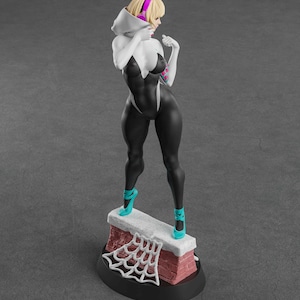 Op de afbeelding: Een beeldje van een vrouwelijke superheld in een zwart-wit bodysuit, staand op een bakstenen structuur met een spinnenwebontwerp. Ze draagt ​​turkooizen hakken en een witte capuchon met roze koptelefoon. De basis is zwart.