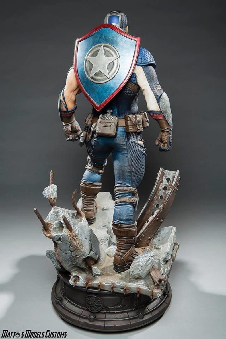 May include: A detailed statue of Captain America from the back, showcasing his iconic shield with a white star on a blue background. He wears a blue and white costume with brown boots and a utility belt. The statue is on a detailed base with a distressed, industrial theme.