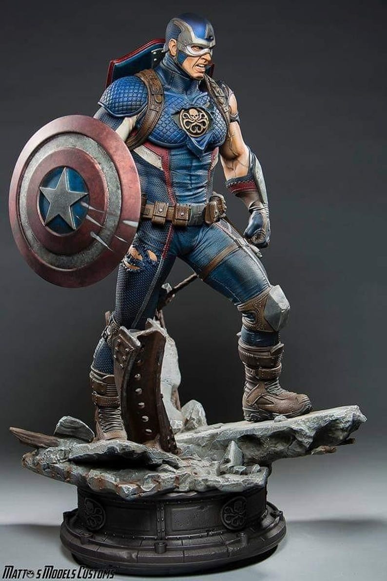 May include: A detailed statue of Captain America, posed with his shield. The figure is in a blue, red, and white costume with fine detailing. The base is a rocky terrain, and the statue has a realistic, muscular physique. The shield has a star.