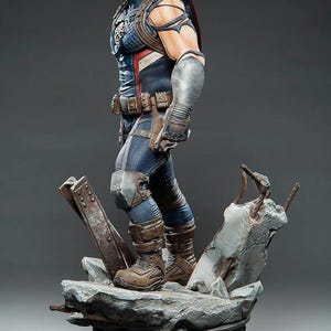 May include: A detailed statue of Captain America in a dynamic pose, wearing a blue, white, and red costume with a shield on his back. The figure stands on a rocky base with metal debris, showcasing intricate details and textures.