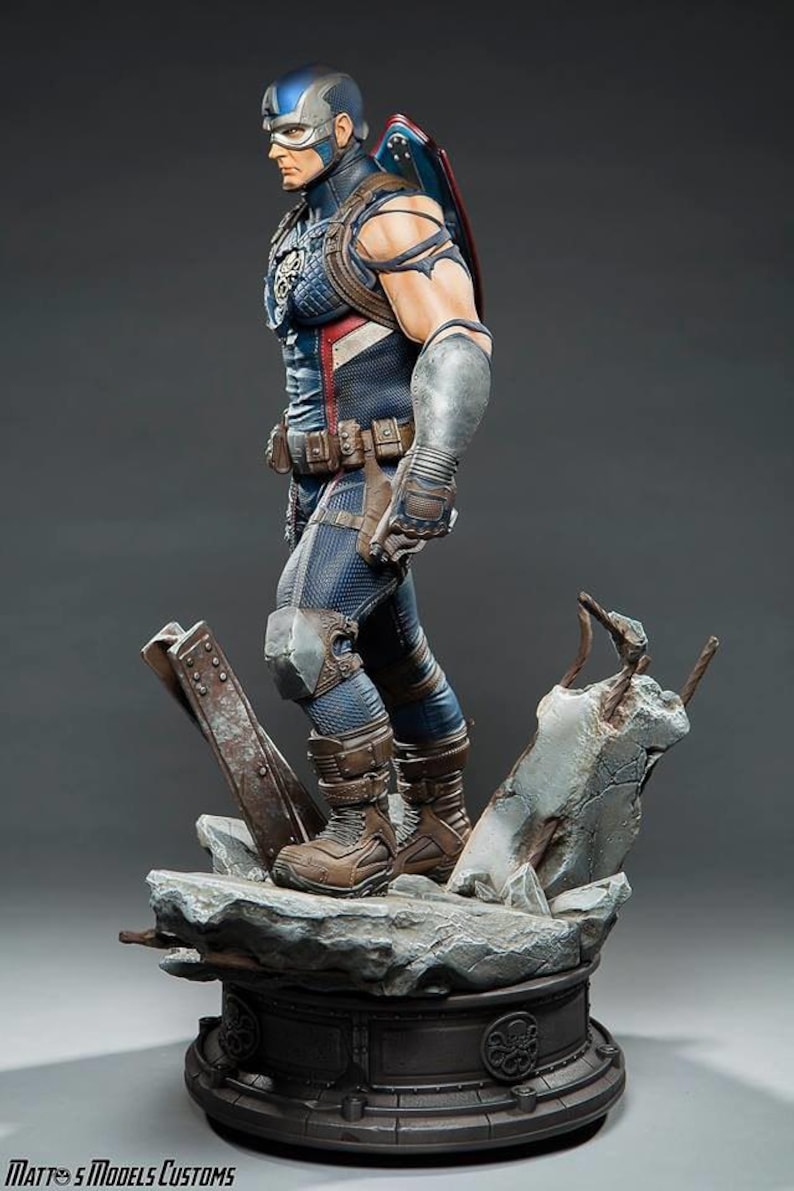 May include: A detailed statue of Captain America in a dynamic pose, wearing a blue, white, and red costume with a shield on his back. The figure stands on a rocky base with metal debris, showcasing intricate details and textures.