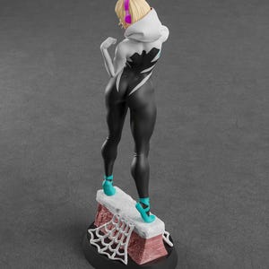 Op de afbeelding: Figuur van een personage in een zwart-wit bodysuit met een spinnenwebontwerp, een witte hoodie en turquoise schoenen. De figuur heeft blond haar en een roze koptelefoon. De basis heeft een bakstenen ontwerp en een wit spinnenweb.