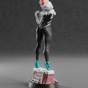 Op de afbeelding: 3D-geprinte figuur van een vrouwelijke superheld in een zwart-wit pak met een spinnenwebontwerp. De figuur heeft blond haar, een witte capuchon en turquoise hakken. De figuur staat op een bakstenen basis.