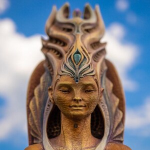 Può includere: Una scultura in legno di una persona con un copricapo dettagliato. La scultura è dipinta in tonalità dorate, marroni e verdi. La persona ha gli occhi chiusi e un'espressione serena.