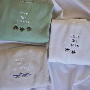Op de afbeelding: Drie sweatshirts met geborduurde berichten en afbeeldingen. De eerste sweatshirt is groen met de tekst "save the turtles" en drie schildpadden. De tweede sweatshirt is wit met de tekst "save the bees" en twee bijen. De derde sweatshirt is beige met de tekst "save the whales" en twee walvissen.