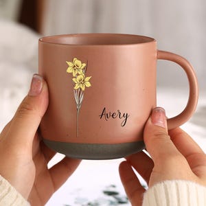 Könnte beinhalten: Eine braune Keramiktasse mit einem gelben Narzissen-Blumendesign und dem Namen "Avery" in schwarzer Schrift. Die Tasse hat einen dunkelgrauen Boden und einen gebogenen Henkel. Die Tasse wird von einer Person gehalten, die einen weißen Pullover trägt.