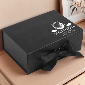 Puede incluir: Una caja de regalo negra con una cinta negra y el texto blanco "PATRIZIA 08.10.2025 BEST GOLFER". La caja rectangular, con un diseño de golf, está sobre una superficie de madera.