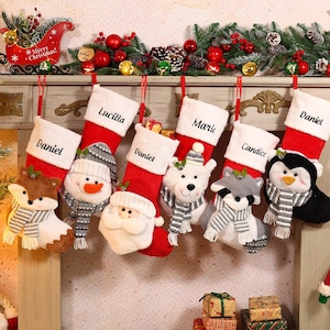 Puede incluir: Una colección de calcetines navideños personalizados en rojo y blanco, cada uno con un nombre bordado en el puño. Adornos decorativos de animales de peluche, como un zorro, un muñeco de nieve, Papá Noel, un oso polar, un mapache y un pingüino, cuelgan de los calcetines.