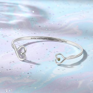 May include: A silver-colored bangle bracelet with a heart-shaped design at each end. The bracelet has the inscription "Mother and Daughter Forever Linked Together." One heart has a clear stone, and the other has a green stone.