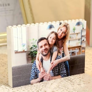Könnte beinhalten: Ein rechteckiger Fotoblock mit einem Familienporträt. Das Bild zeigt einen Mann, eine Frau und ein Kind, die lächeln. Der Block ist weiß mit einer strukturierten Oberseite, die an Spielbausteine erinnert. Ein dekorativer Bilderrahmen und Bücher befinden sich im Hintergrund.