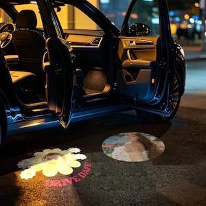 May include: A black car with an open door projecting two circular light displays onto the ground. One projection shows a person in a floral top, while the other features a yellow flower graphic with a photo and the red text "DRIVE SAFE".