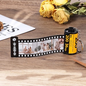 Pode incluir: Um porta-chaves de cartucho de filme de 35 mm amarelo e preto com uma tira de filme anexada apresentando fotografias impressas de uma família e um ícone de código musical. O cartucho tem texto incluindo 200, 36 exp e Kodak 200 36 process C-41.