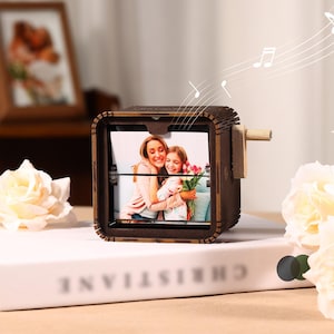 May include: A dark brown wooden music box featuring a square frame that displays a photograph of two people embracing while holding a bouquet of tulips. The box has a light-colored wooden hand crank on the side and a slotted top design.
