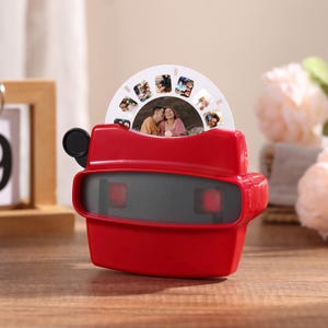 May include: A bright red plastic stereoscope viewer featuring a circular white photo reel inserted into the top slot. The device has two rectangular viewing lenses on the front and a black circular lever on the left side for advancing the images.