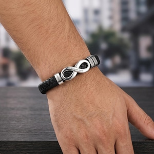 Könnte beinhalten: Schwarzes geflochtenes Lederarmband mit einem silbernen Unendlichkeitssymbol und personalisierten Namensschildern. Das Unendlichkeitssymbol ist mit den Worten "Forever Love" graviert. Ein stilvolles Accessoire.