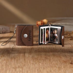 Puede incluir: Un mini álbum de fotos de cuero marrón con la palabra "Daddy" grabada en la parte delantera. El álbum se cierra con un broche y tiene una pequeña correa para la muñeca. El álbum está abierto, revelando varias fotos pequeñas.