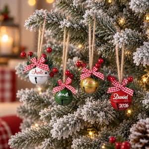 May include: Four round metal jingle bell ornaments hanging from twine. The bells are white, green, gold, and red, each featuring a red and white gingham bow, red berry sprigs, and personalized names including Kate, Janet, Paul, and Daniel with 2026.