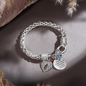 May include: Silver-toned bracelet with a braided chain design. The bracelet features charms including a pair of wings, a heart-shaped clasp, and a circular charm with the text "A sister is God's way of making sure we never walk alone". Red and blue gemstones are also present.