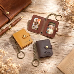 May include: Three small leather-like photo album keychains with metal chains and rings. One is open, displaying two color photographs of people, while the others are closed. The yellow one is engraved with "Julian" and a heart, and the brown one with "Dad".