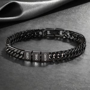 May include: A black stainless steel curb chain bracelet. The bracelet features three rectangular nameplates with the names "CARTER", "DANIEL", and "ALVARO" engraved in white. The bracelet is secured with a clasp.