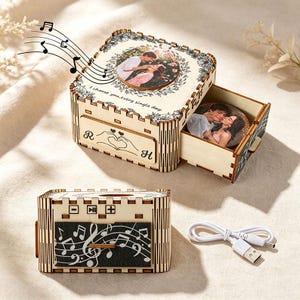 May include: A square wooden music box with a light wood finish, featuring a circular photo of a couple on the lid and the text I choose you, every single day. The front shows the letters R and H with a heart-shaped hand gesture. A white USB cable is included.