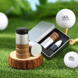 May include: A golf ball marking kit with a silver and wood-tone stamp, ink bottle, and golf balls. The golf balls have the text "Jonny M" printed on them. The kit is presented on a green surface, suggesting a golf course setting.