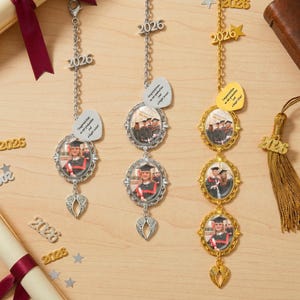 Może przedstawiać: Trzy zawieszki do samochodu z motywem ukończenia szkoły. Dwie są w kolorze srebrnym, a jedna w kolorze złotym. Każda zawieszka zawiera zdjęcie, rok 2026 i etykietę w kształcie serca z tekstem. Zawieszki mają ozdobne akcenty skrzydeł.