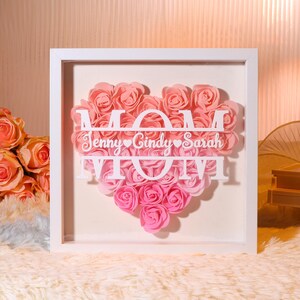 Op de afbeelding: Witte ingelijste schaduwdoos met een roze en wit ombre hart gemaakt van papieren rozen. Het hart heeft het woord "MOM" uitgesneden met de namen "Jenny, Cindy, Sarah" eronder geschreven.