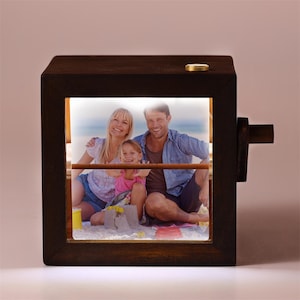 May include: A dark brown wooden photo cube with a family photo displayed inside. The cube has a small gold-colored button on top and a wooden handle on the side. The family photo shows a smiling couple and a child.