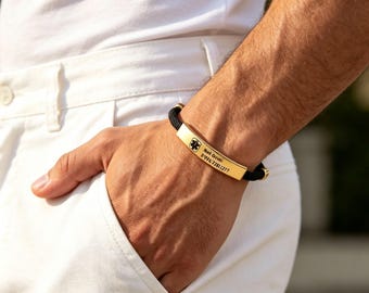 Puede incluir: Una pulsera de alerta médica con una banda de cordón negro y una placa de identificación de metal dorado. La placa está grabada con un símbolo médico de caduceo negro, el nombre "Nok Demi" y la secuencia numérica "091673811277" en letra clara.