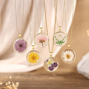 Puede incluir: Seis collares dorados con colgantes circulares, cada uno con una flor prensada diferente preservada en resina transparente. Las flores incluyen un crisantemo morado, una margarita amarilla, una flor rosa, una blanca, un pensamiento y una margarita blanca.