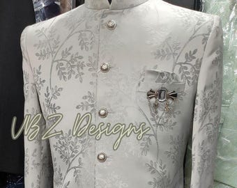 Puede incluir: Chaqueta de traje gris claro con un estampado floral y botones decorativos. La chaqueta tiene cuello mandarín y un pañuelo de bolsillo con un alfiler decorativo. El texto "VBZ Designs" es visible en la chaqueta.